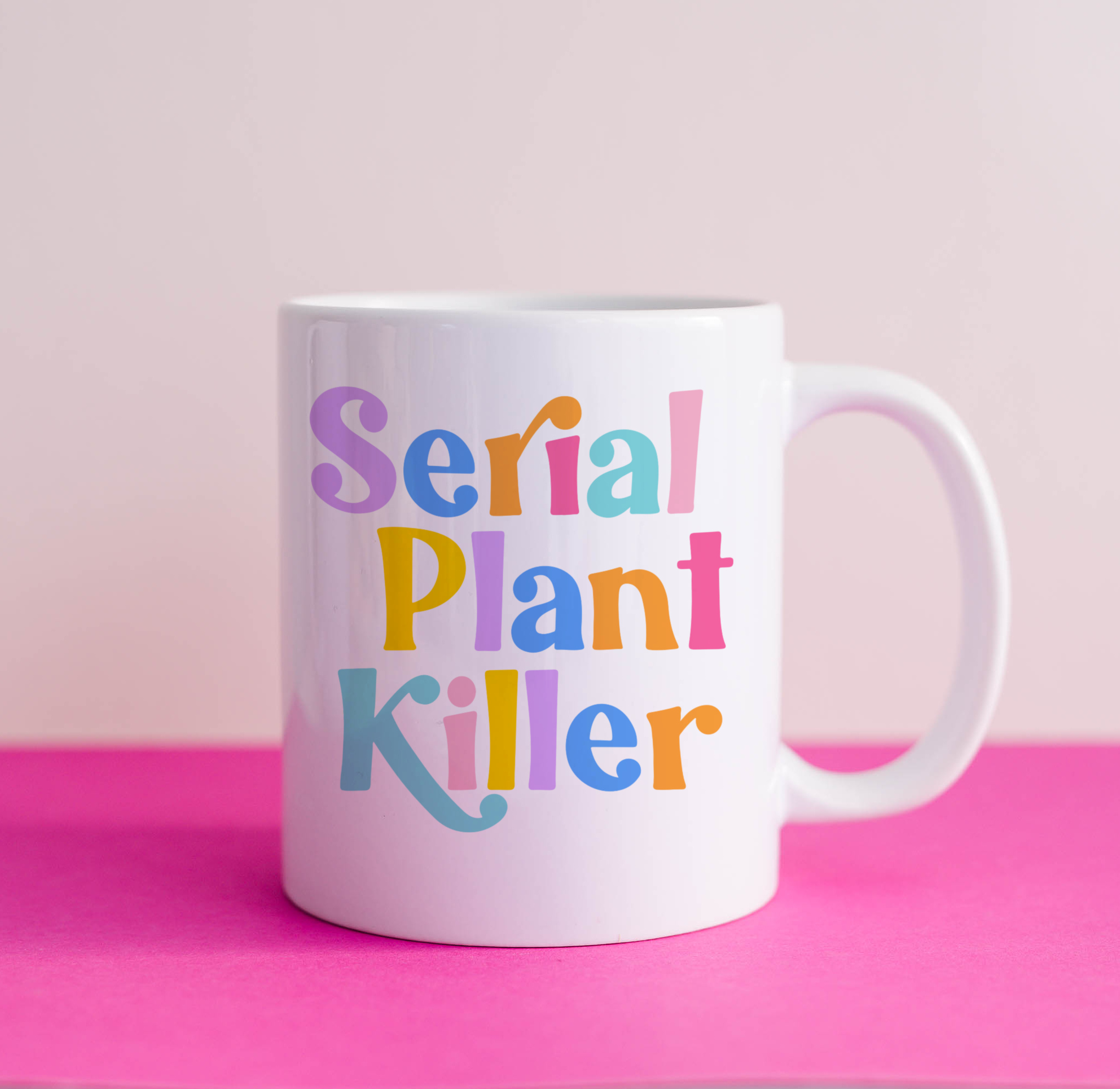 "Studio shot of 'Serial Plant Killer' mug against pink backdrop. Vibrant design with rainbow-colored text."
